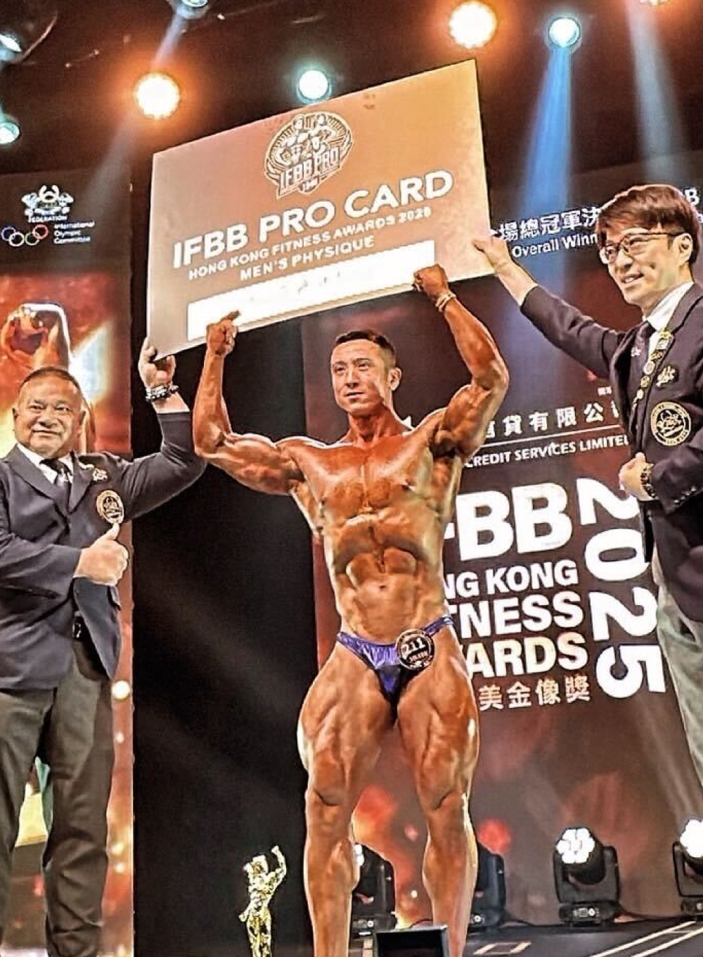 ステージ上でポーズをとるボディビルディングのコンテスト参加者。青いショートパンツを履き、左足に「54」の番号が表示されている。