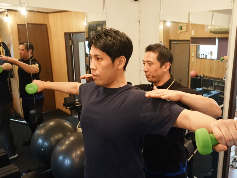 トレーナーが男性にダンベルを使った肩のエクササイズを指導している。
