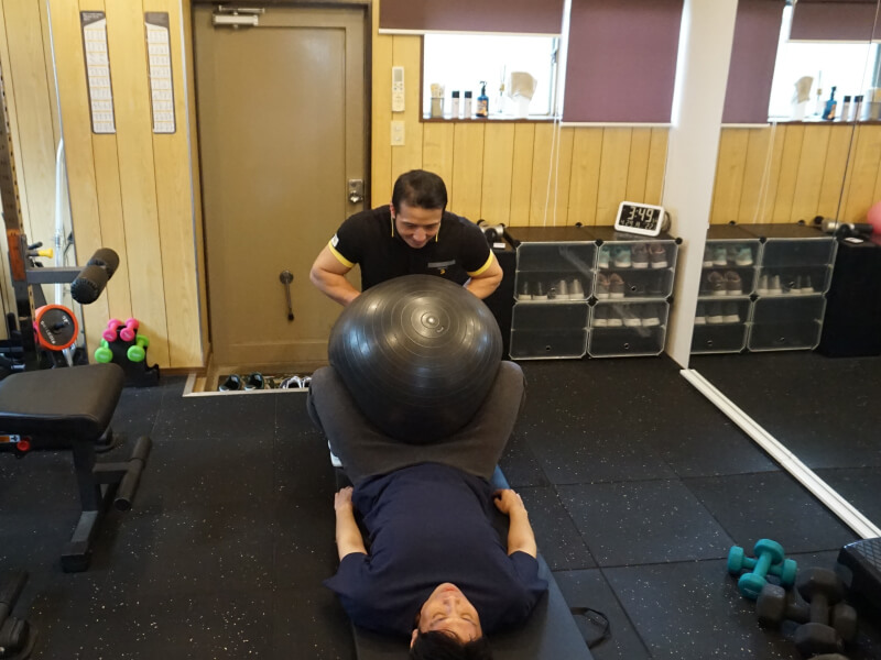 トレーナーが男性にバランスボールを使ったエクササイズを指導している。